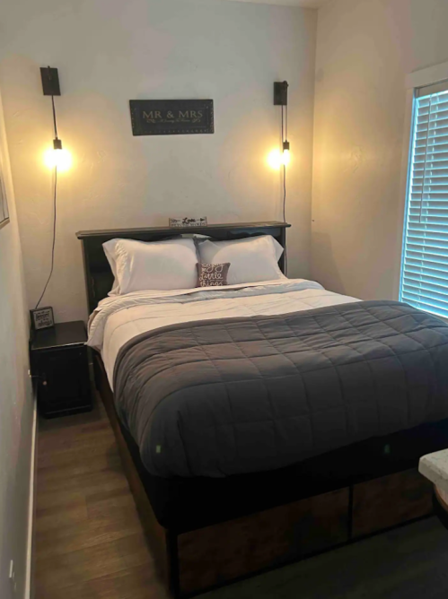 Cozy bedroom featuring a bed with gray bedding, bedside lamps, and a "Mr. & Mrs." sign on the wall.